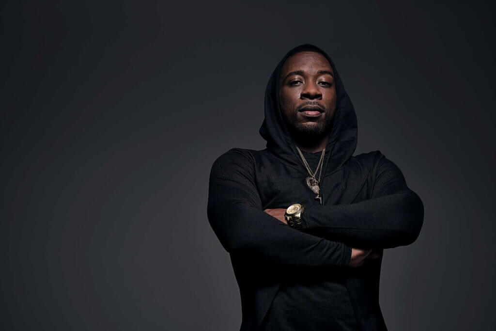Iquail, a dark, brown-skinned man with short, cropped cut hair and a goatee, stands in dark lighting with his arms folded across his chest while wearing a black hoodie shirt, a gold watch, and gold chains.