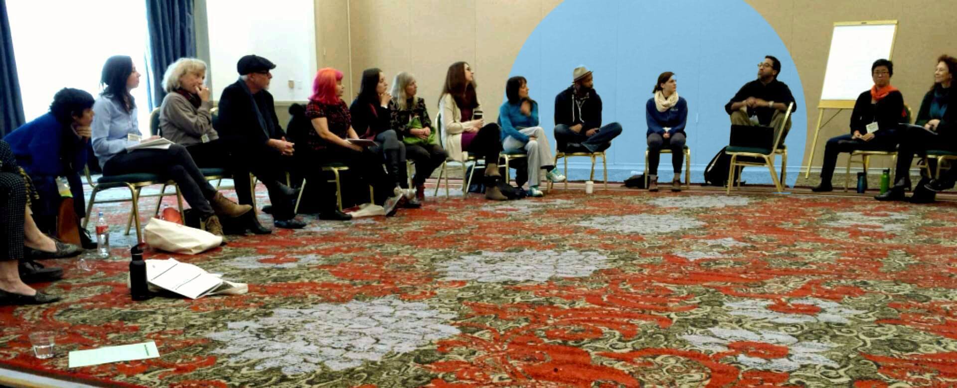 A diverse group of people are seated in a wide circle of chairs in a conference or workshop setting, engaged in conversation. The room has a patterned red and green carpet, and a blue circle shape is edited onto the back wall behind a flip chart easel. Some attendees are taking notes while others listen attentively.