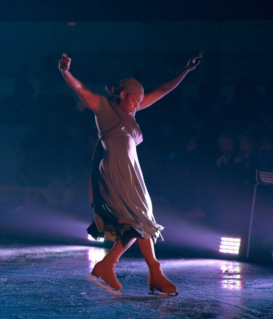 A Black woman in a gray dress with a full skirt wearing a matching gray head wrap in figure skates on the ice with both arms raised performing a movement that brings her right skate in front of her left skate which is balanced on the toe pick. Her gaze is facing down toward the ice.