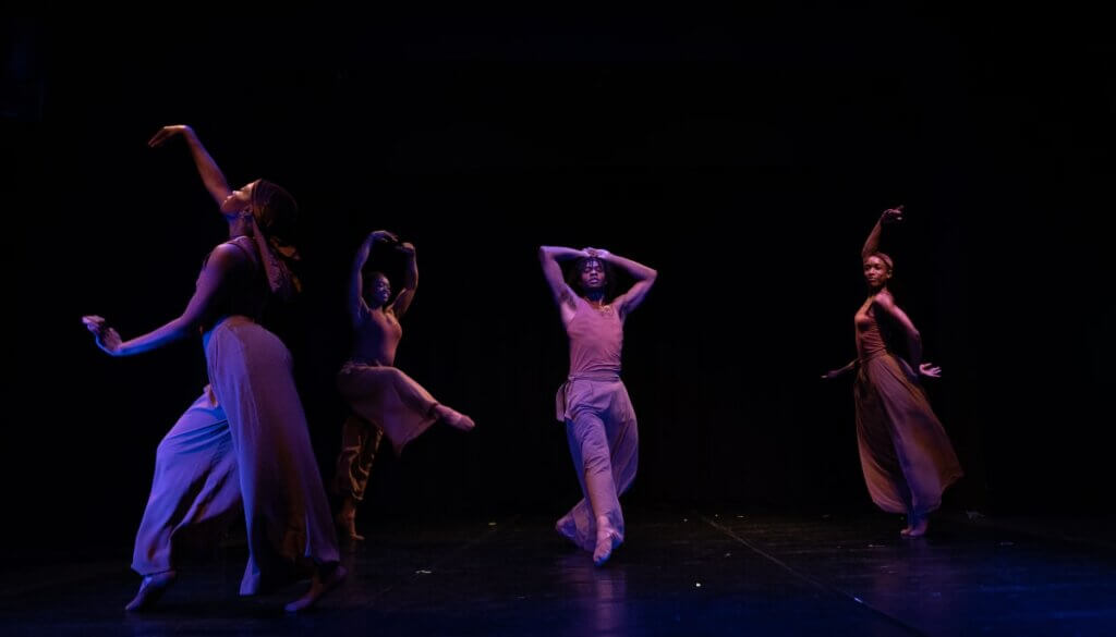 Four brown-skinned dancers of varying builds, dressed in close fitting brown tank tops, brown flowing pants, and flesh tone ballet shoes are spread out across a dark stage, captured in mid-motion. The dancers make varying shapes with their bodies, raising their arms above their heads, rising on the balls of their feet, and reaching with their legs.