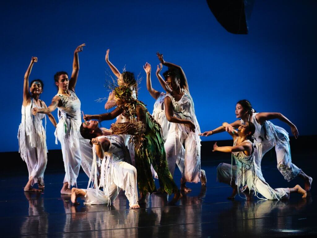 Against a lit, blue background, five femmes in shredded, white costumes stand with open arms, surrounding one woman who kneels in a backbend. A non-binary artist in green wearing a crown of golden twigs paints the kneeling woman’s forehead. To the right, one woman kneels, facing the rest, with arms rounded, her sternum opening to the group. Another woman leans over her and reaches toward the group. All the artists are of various medium-to-dark brown skin tones.