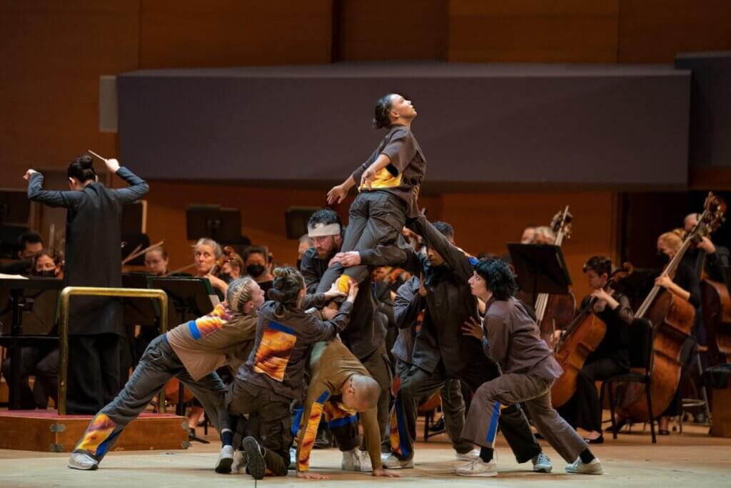 Dancers are on stage in front of an orchestra. A Sicilian American female facing stage left, looking up with arms extended is standing on the back of Vietnamese American male, bent at waist with hands on the floor, head facing the audience. From behind, standing Caucasian female holds Sicilian dancer’s left leg, Chinese American female kneeling, holds right leg. Sicilian dancer’s thighs are supported by standing Caucasian male and her torso is supported by standing Mexican American male extending his arms to her stomach. He is supported on both sides by Romanian-Jewish American female and Colombian/African American female.