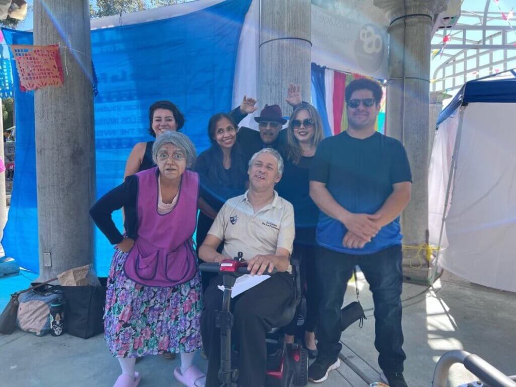 An outdoor space with two pillars in the back and 7 people standing in front. Hanging to the left is a blue tarp, and to the right are colored decorations. The back row from the left, a young light-skinned woman with dark hair, a brown skinned woman with dark hair, a light-skinned man with sunglasses and a red hat waving both hands, a light-skinned woman with sunglasses and blondish dark hair, a light-skinned male with dark fluffy hair. In front from the left, a woman dressed as an older lady and a brown-light skinned man on a mobile scooter.