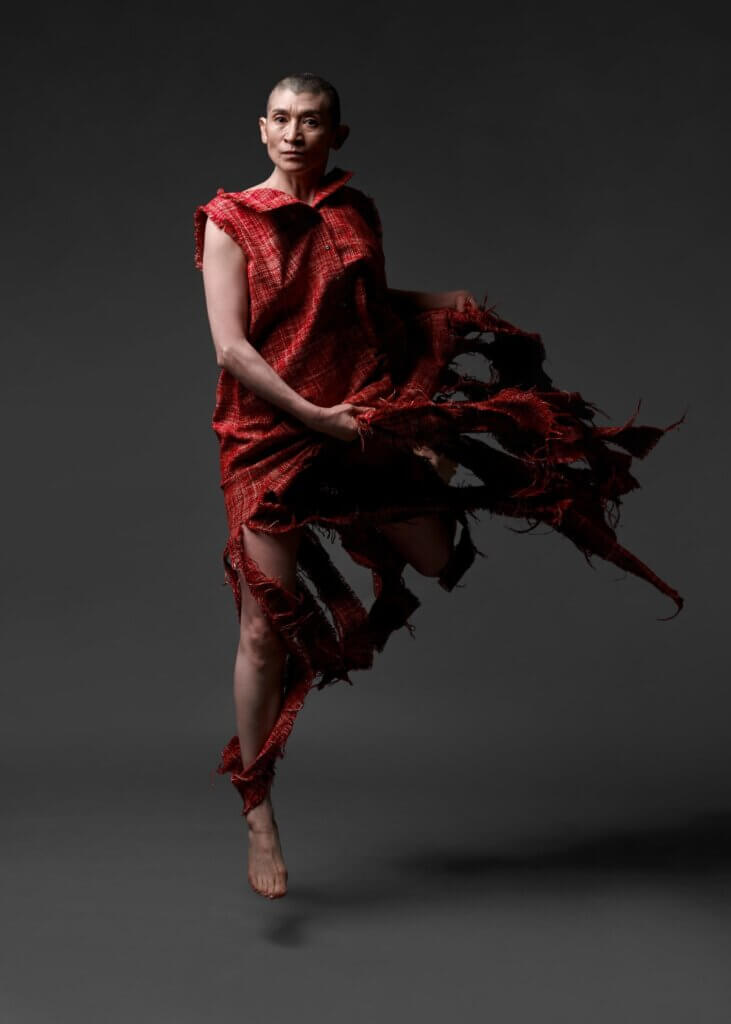 A queer, androgynous, medium-skinned Mexican woman with a shaved head, dressed in a tattered, red, sleeveless, sack-like garment, jumps straight up on one leg while staring intensely into the camera in a vacant dark gray space. A shadow of this figure can be seen on the gray flooring.