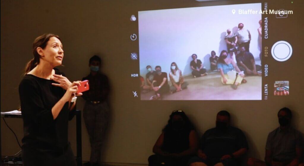 A light-skinned Latina woman with long brown hair tied in a ponytail and wearing a black dress holds a red iPhone towards the audience as she sings. The image of the audience that the iPhone is recording is projected behind her. Many of the audience members are wearing masks. Other audience members are visible sitting below the projected video.