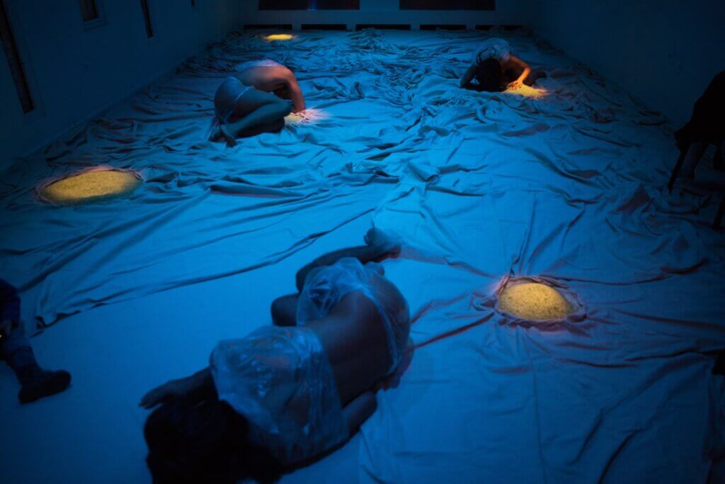 Three androgynous dancers are sprawled on the floor of a blue-lit room whose floor is covered by crumpled sheets with circles of sand appearing here and there and lit by golden lights. The room is long, with covered windows along the left side wall and back wall.