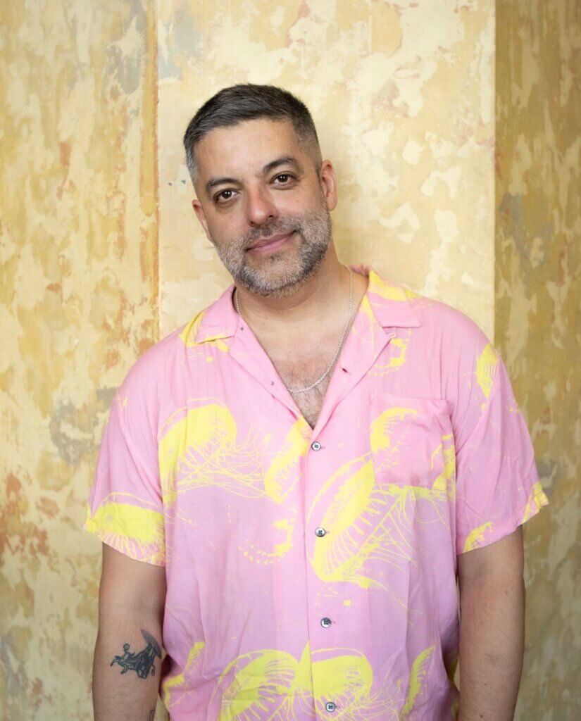 Miguel Gutierrez, a light brown Latino man, leans against a dappled yellow wall. He has short dark hair and a short grey beard. His head is slightly tilted, and he is smiling. Miguel is wearing a silver chain necklace and a pink shirt with electric yellow jellyfish printed on it. On his arm is a Taurean bull tattoo.