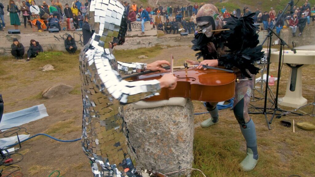 A performer wearing a reflective bodysuit moves a bow across a horizontally positioned cello while a second performer wearing a costume consisting of a camouflaged bodysuit with a black feather cape and silver face paint with a black line through the eyes plucks at the same cello. A large crowd looks on in the background.