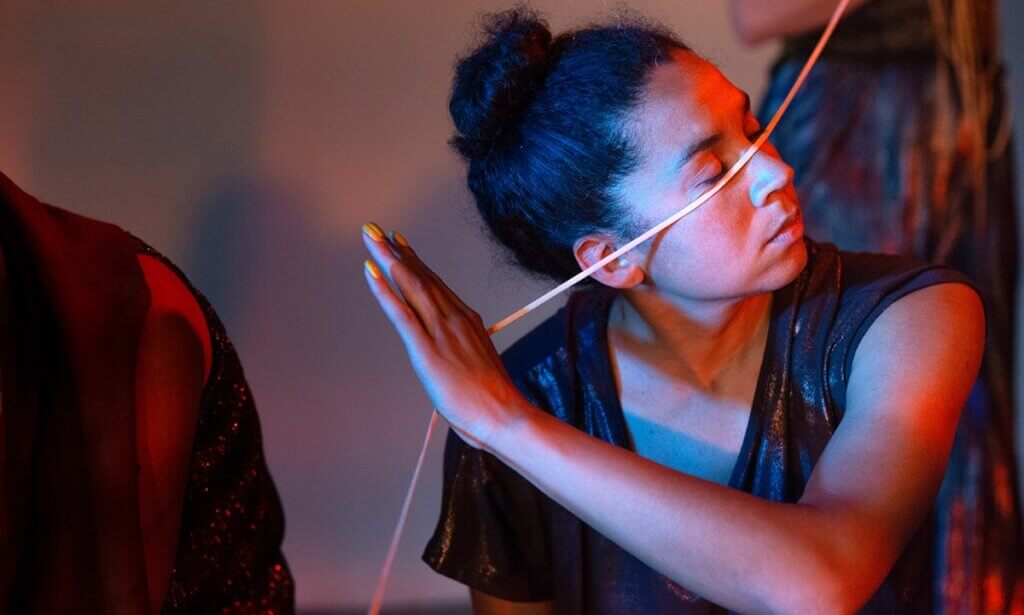 The photo features Lela Pierce, a Black and multiracial woman with brown skin and curly shoulder-length black hair. She drapes a pale, thin deerskin lace under her closed eyes and across the bridge of her nose. She reaches for an open hand with yellow fingernails across her to support the deerskin as it travels past her inclined head. Others move around her.