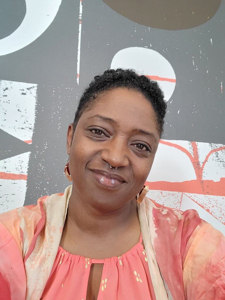 A headshot of a medium-skinned African American woman with short, black, curly hair, smiling and looking into the camera. She wears a peach and light tan colored shirt that matches a gray and peach colored background. She is wearing a septum nose piercing and earrings that feature wood and copper.