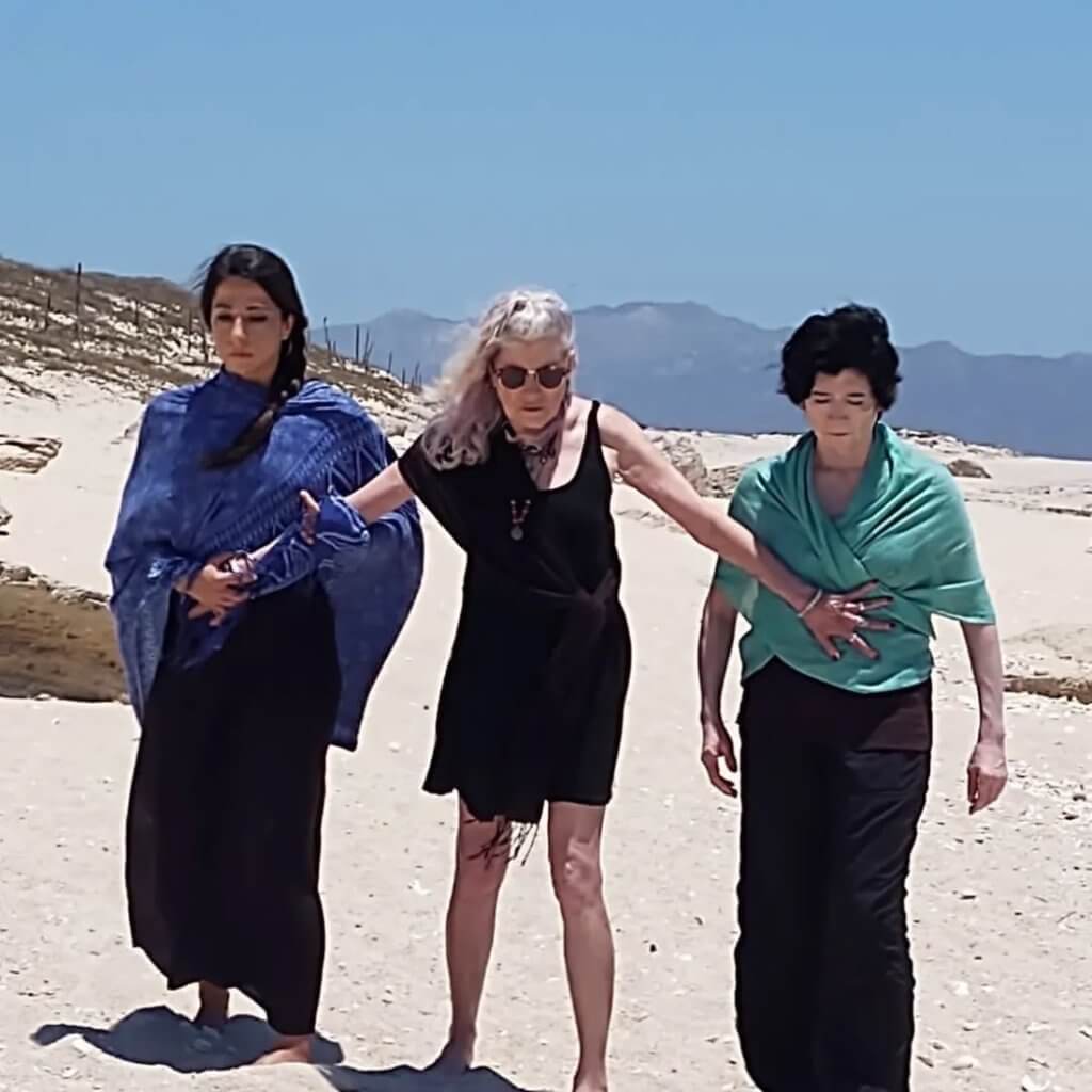 Three performers on a beach, with sand dunes and mountains in the distance. A central figure with light hair in a black dress and sunglasses reached out to stop the other two, who are on either side of her and both have dark hair and are wearing black with blue and aqua rebozo shawls, from moving forward. All three look down at the sand in the mid-day sun.