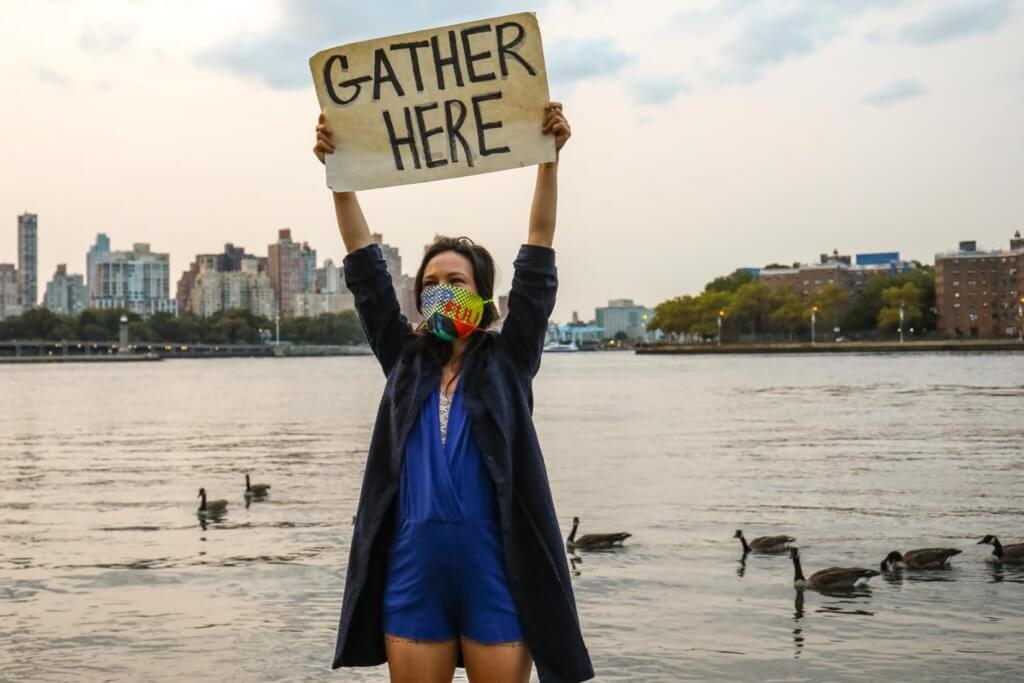 Emily Johnson, a Yup’ik woman, stands at the edge of the East River in Lenapehoking. She has long black hair and wears a vibrant face mask designed by artist Jeffrey Gibson, with the words “Power” and
“Full” on it. Her arms are raised over her head, and she holds up a hand-written sign that reads “Gather
Here.” Behind her, the gray water is spotted with ducks, and some of the Manahatta skyline is visible.