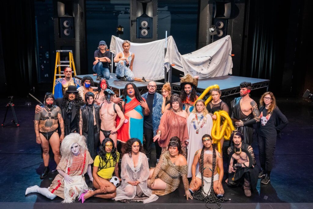 Photo of film cast and crew, roughly 30 people, dressed in vibrant and elaborate costumes. Some have more elaborate costumes with gold touches; some wear simple tunics with blue touches on their face make up; and some are in mostly black with red makeup. They stand and kneel in front of a stage.