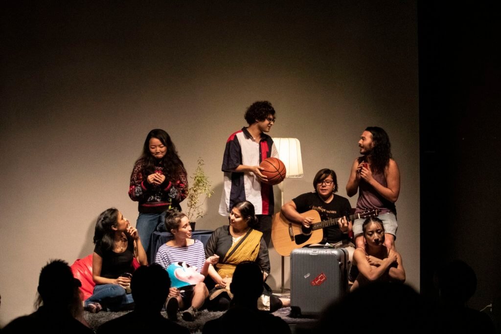8 artists of Pan-Asian descent are gathered on a small platform in front of the audience. The performers are sitting and standing. They wear casual clothes and are talking to one another. One person plays a guitar, and one person is huddled in a corner by a suitcase.
