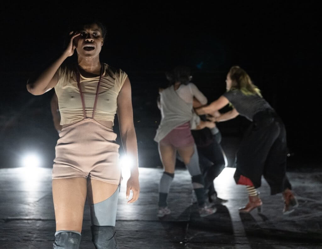 A distressed young Black woman singing onstage in foreground. Three people in the background tackle a fourth person.