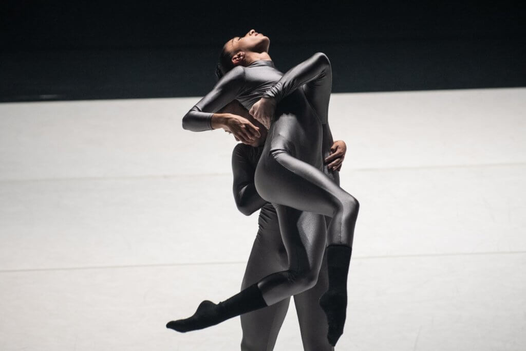 An African American person and Greek-Indian person support each other in a suspended arch lift wearing shiny gray one piece suits. They are lit in stark light.