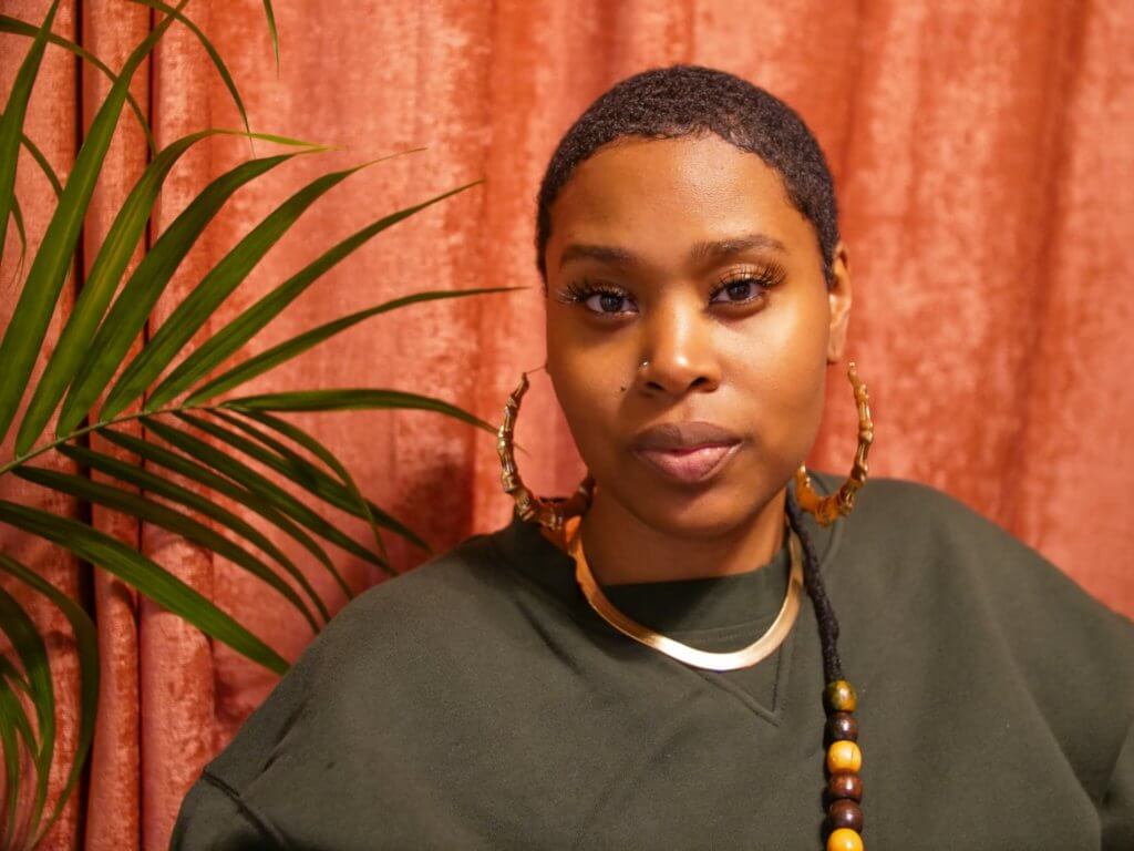 A medium-dark skinned woman with a short black hair cut looking straight ahead with a dark green long sleeve shirt, gold hoop earrings and a loc with beads at the end. Her background is a salmon color with a green palm tree on her right side.