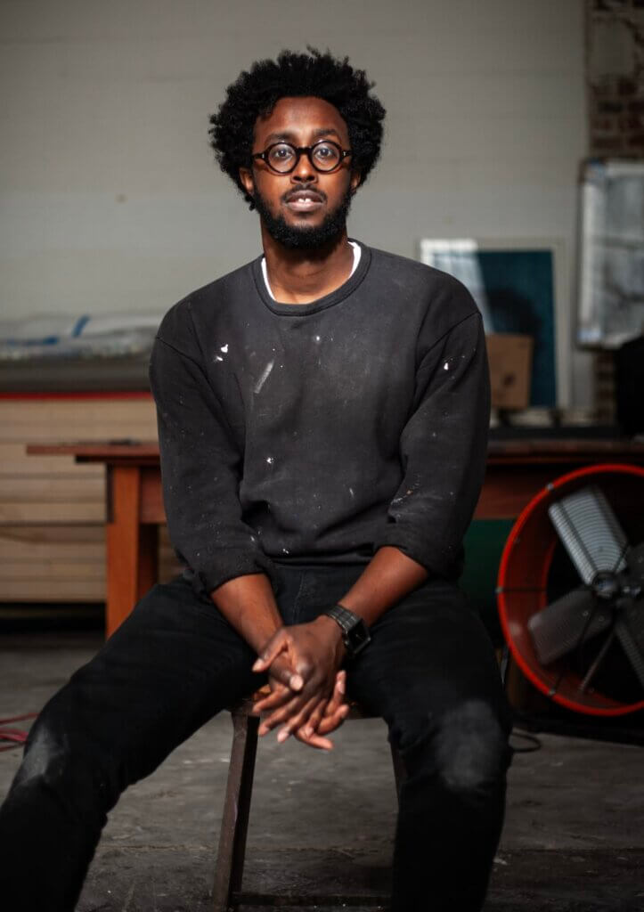 Abdi Farah An image of a dark-skinned African American man wearing black-rimmed eyeglasses, a black sweatshirt, and black pants–looks at the camera while sitting on a stool in their studio.
