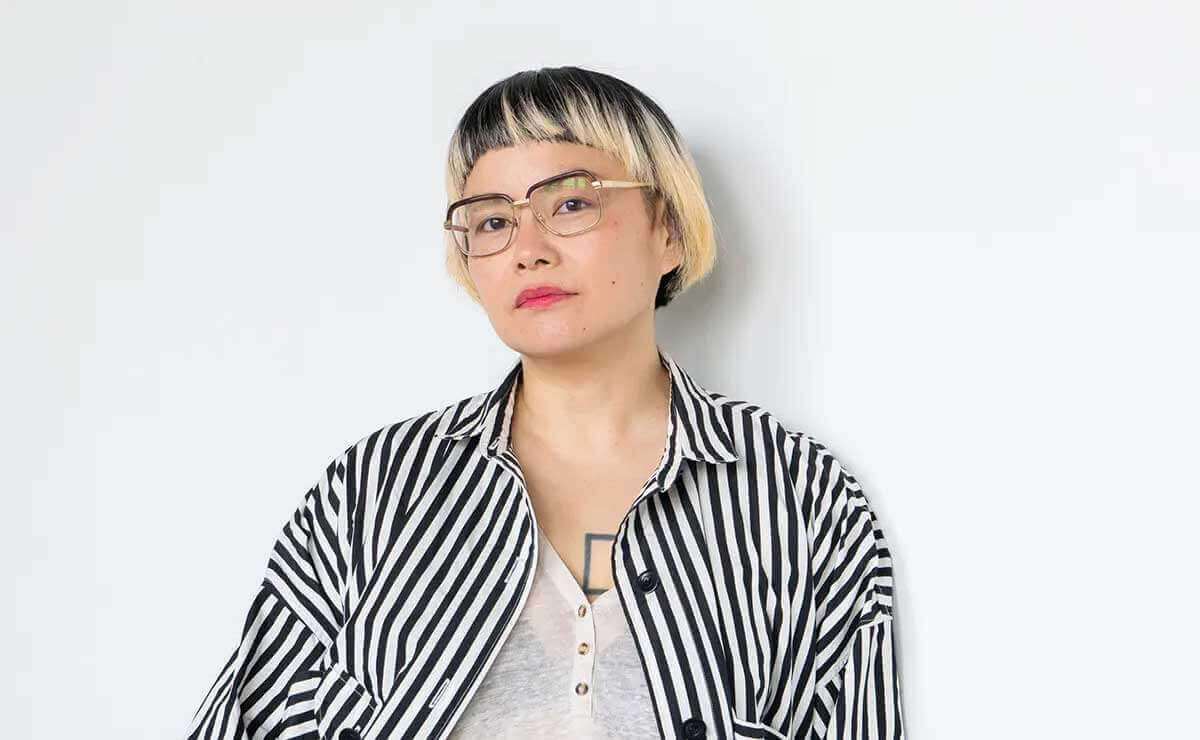 Headshot of Anne Ishii, an Asian woman standing against a plain white background, wearing a matching black and white striped outfit, including a loose-fitting top and pants. She has light skin and short, straight hair with dark roots and blonde ends. She is shown from the chest up, wearing glasses and has a neutral expression, with the bold striped clothing being a focal point against the minimal backdrop.