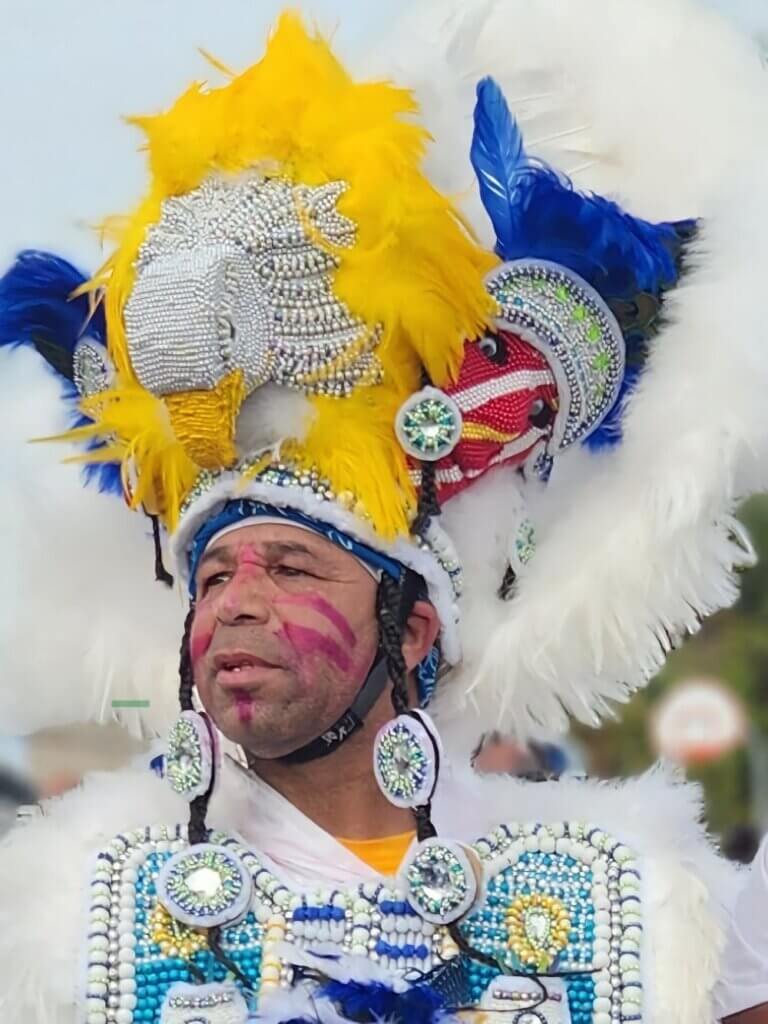 Anthony Hingle Jr. | Original Spy Boy of the Black Feather Tribe An image of a medium-skinned man looking off into the distance wearing a large white, yellow, and blue feathered headpiece with the head of a white eagle atop it. He has red paint on his face and wears a white and blue beaded and feathered Black Indian suit.