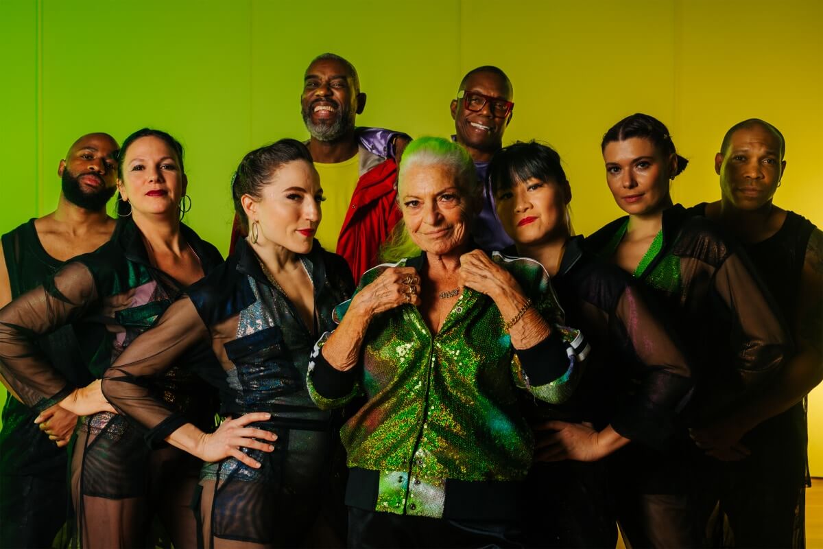 A multi-racial, multi-gendered group of people look out at the camera with smiling face in front of a green background.