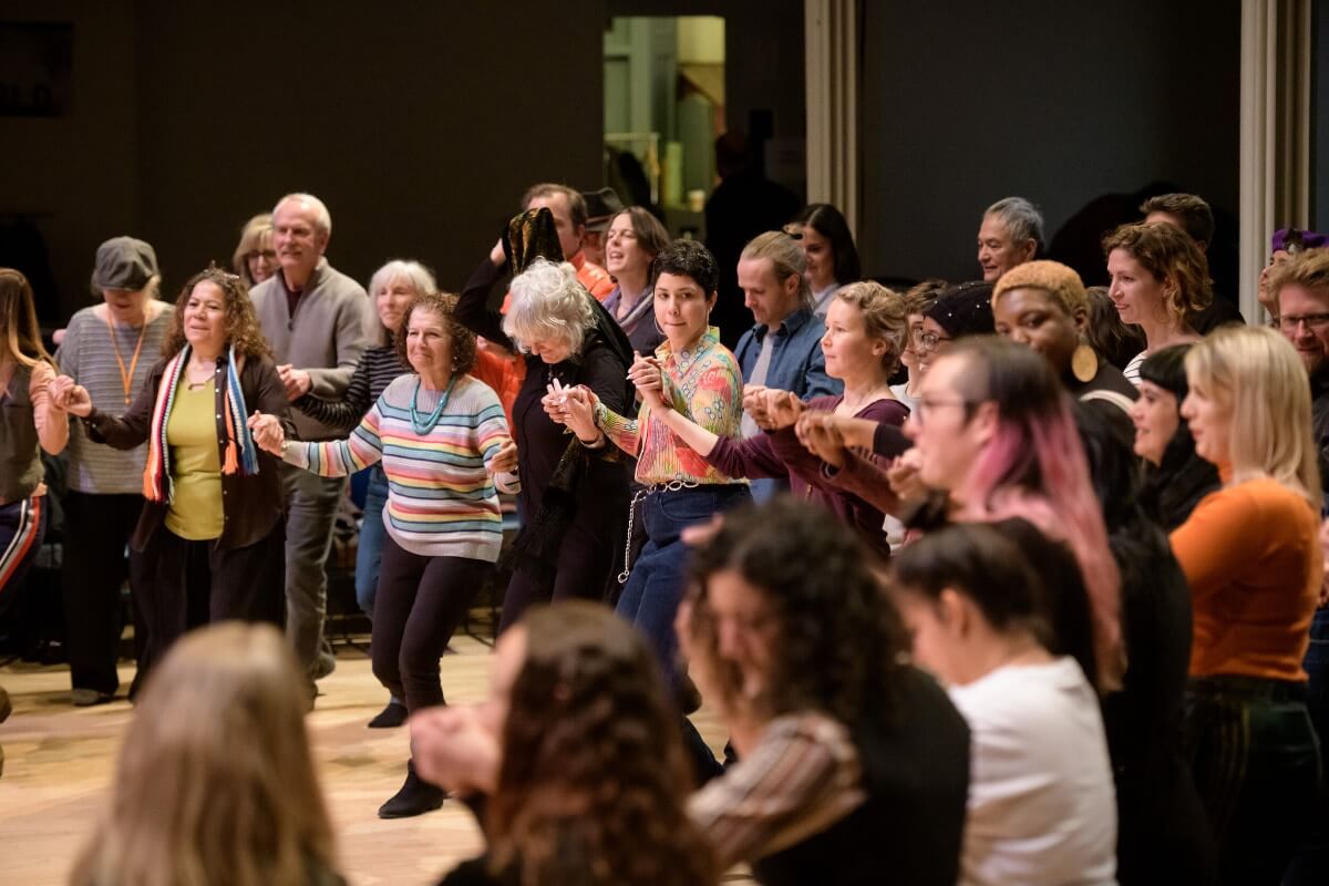 A large groups of people are doing a dance together facing the same way on a wooden floor.
