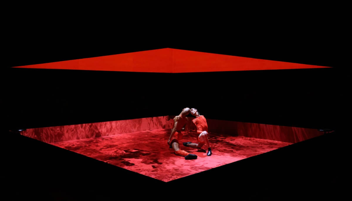 apap 24 - shamel touch of red 2 men of color lunge towards each other, heads almost touching; feet almost touching. They are both dressed in red tight-fitted athletic/wrestling clothing while inside of a red boxed space. The floor displays a red moving lake, while the interior of the box is engulfed in a striking deep red color. The outside of the boxed ring is black with the middle cut out which functions as the viewing point into the red box.