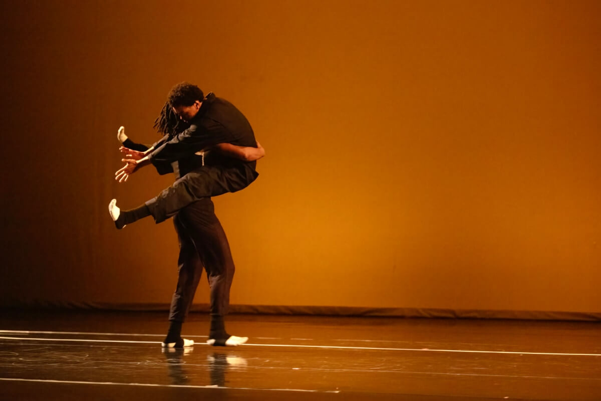 Two black men stand in an embrace on stage right. One man holds the other in a koala hug. The man being held has his arms and legs outstretched beyond his partner's body. They stand in front of an orange background. They both wear black boilersuits and black and white socks.