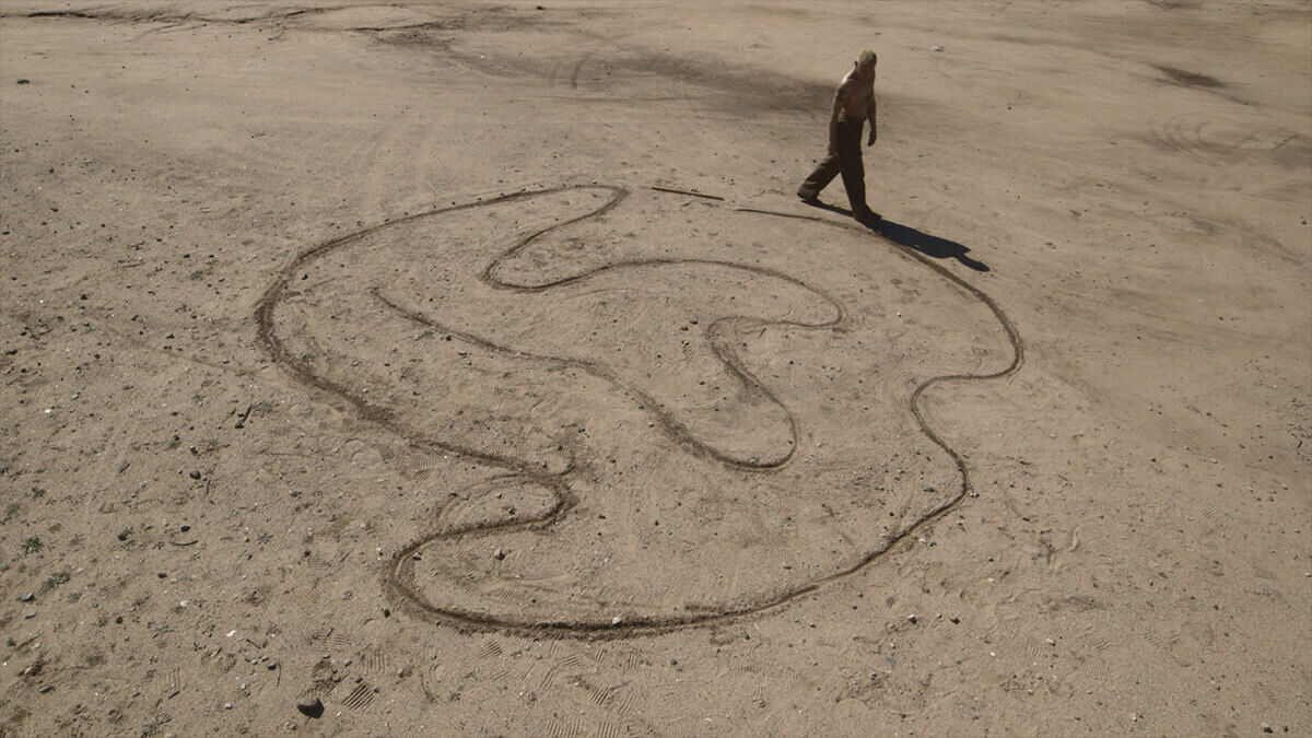 A person bathed in sunlight walks around a sacred shape carved in a brown dirt ground.