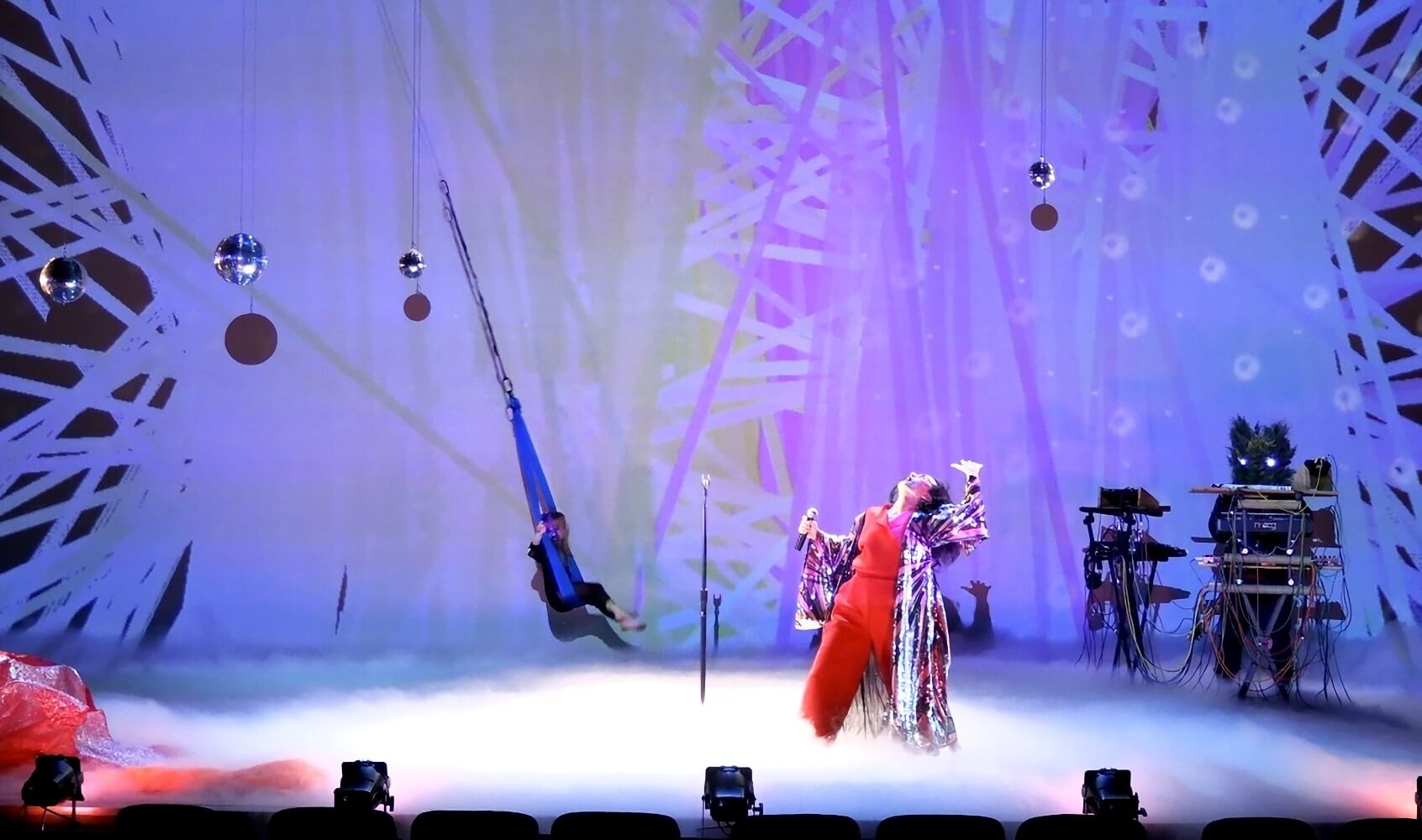 On a brightly lit, fog-covered stage, against a backdrop of purple, lavender, and pale blue lines crossing at random angles so that they form a nearly solid mass, a performer in a red pant and matching red shirt, and wearing a purple, white and red flower patterned robe, poses with one leg stepping forward and head thrown back . In the background, far upstage, another figure in black sits in a blue fabric harness and swings. Four disco balls hang above the performers. On the far right side of the stage is a DJ table with cables and equipment.