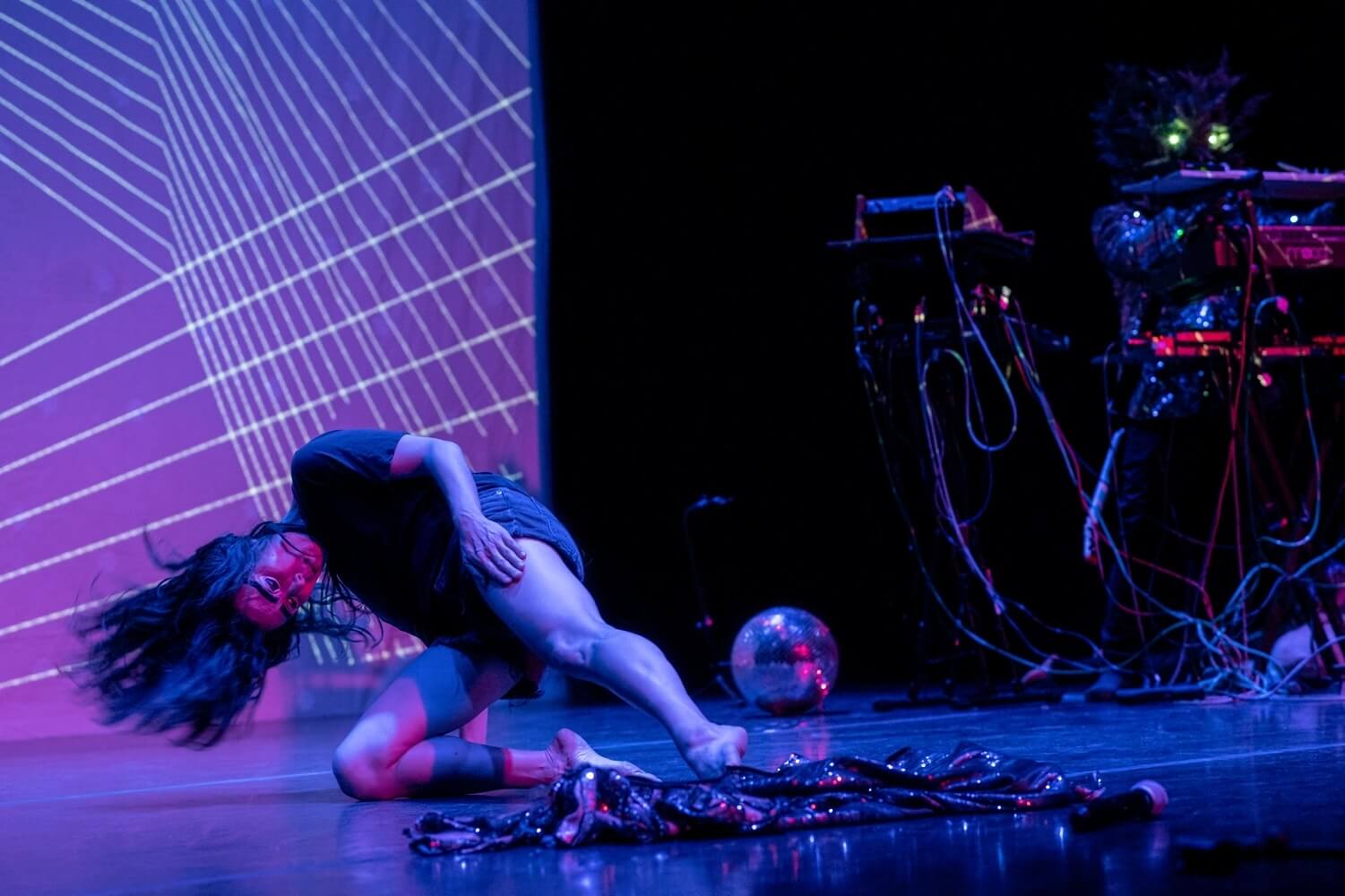 On a stage lit in purple and blue, a female-presenting dancer in a short-sleeved dark shirt and dark shorts presses a knee onto the ground and extends the other leg, while tilting her head down and swinging it so that her long black hair flies out away from her skull. In front of her is a rumpled and highly reflective swatch of fabric, and behind her a disco ball rests on the ground. The left side of the background shows part of a backdrop for the performance, which is a series of sets of thin white lines crossing over each other like string art. On the right side we can see part of what appears to be a DJ table with cables and equipment.