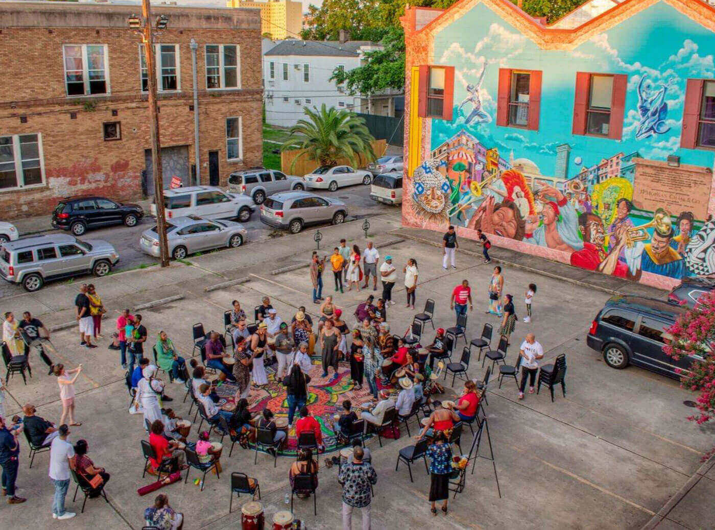 A vibrant community gathering takes place in the parking lot outside the Ashé Cultural Arts Center, with people seated in a large circle, many playing drums and dancing in the center. The background features a colorful mural painted on the building’s wall, depicting musicians, cultural icons, and scenes of celebration. Onlookers and participants fill the space, creating a joyful, lively atmosphere surrounded by cars and neighborhood buildings.