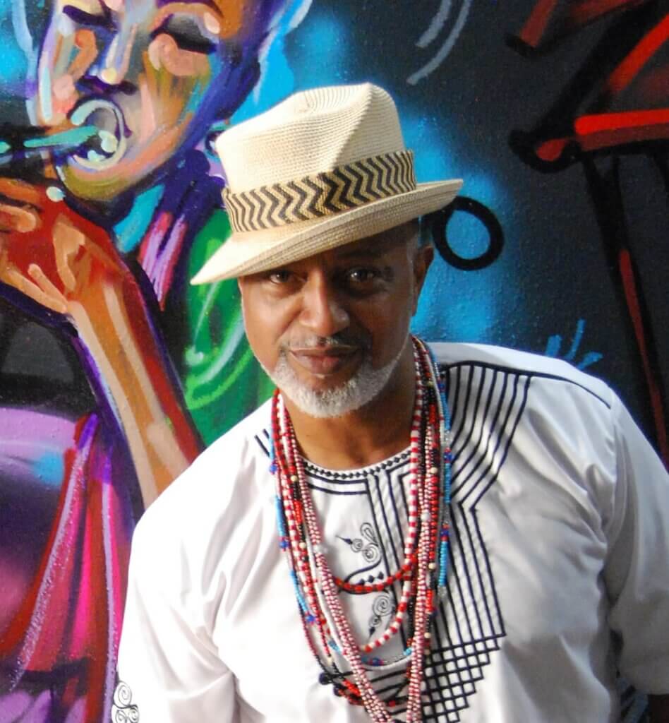 Baba Marcus Ṣàngódoyin Akinlana The image is of a medium, dark-skinned African-American man with a light gray mustache and beard. He is wearing a beige straw fedora, titled to the side, and a white kaftan with navy blue embroidery around the collar. Behind him is a colorful mural featuring a musician playing a horned instrument.