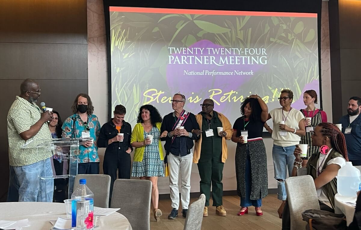 In a large hotel meeting or conference room, ten adults of various skin tones stand in a line in front of a large projection screen. They are all holding white paper cups and wearing lanyards, and otherwise dressed in ordinary everyday contemporary clothing. They are all listening to and looking at a tall black man who is standing to their right--our left--behind a clear acrylic podium and speaking into a microphone as he addresses them. In the foreground on the left is the edge of a large round table covered in a white tablecloth, with three cloth-covered gray chairs arranged haphazardly about it. In the foreground on the right is the edge of another similar table, but on this side there is a black woman sitting in one of the chairs and looking at the man speaking. She is wearing a short-sleeved white t-shirt and black, leather-looking vest, and she has tightly braided black and dark-reddish-brown hair that's pulled back. She is wearing large round pink earrings that feature the continent of Africa. Behind everyone, projected on the screen, is an image of plants and the words, "Twenty Twenty-Four Partner Meeting, National Performance Network." Behind the heads of the row of people, partially obscured, is more text that reads, "Steve is Retiring!"