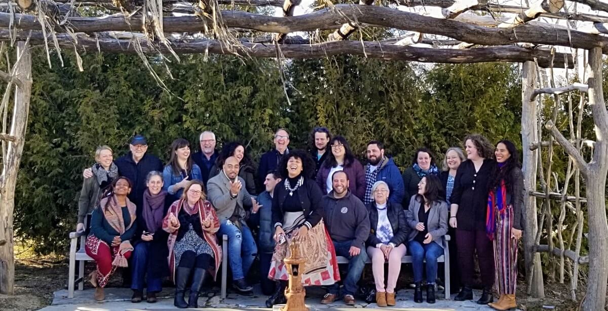 A large, diverse group of nearly two dozen adults are gathered under a large, open rectangular trellis made of untreated tree trunks and branches. The trellis is approximately 15 feet tall, dwarfing the group of people. They are dressed snugly but not in a way that implies it's very cold outside. Immediately behind them is a wall of fir trees.
