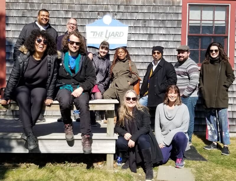 A diverse group of eleven adults pose for a photo on and around an unpainted picnic table in front of building clad in weathered wooden shingles with red wooden framed square windows. There is a blue and white sign on the wall that says "The Yard." They are all dressed snugly but not in a way that implies it's very cold outside. It is quite bright, though, and several of them are wearing sunglasses. They are all smiling or laughing.