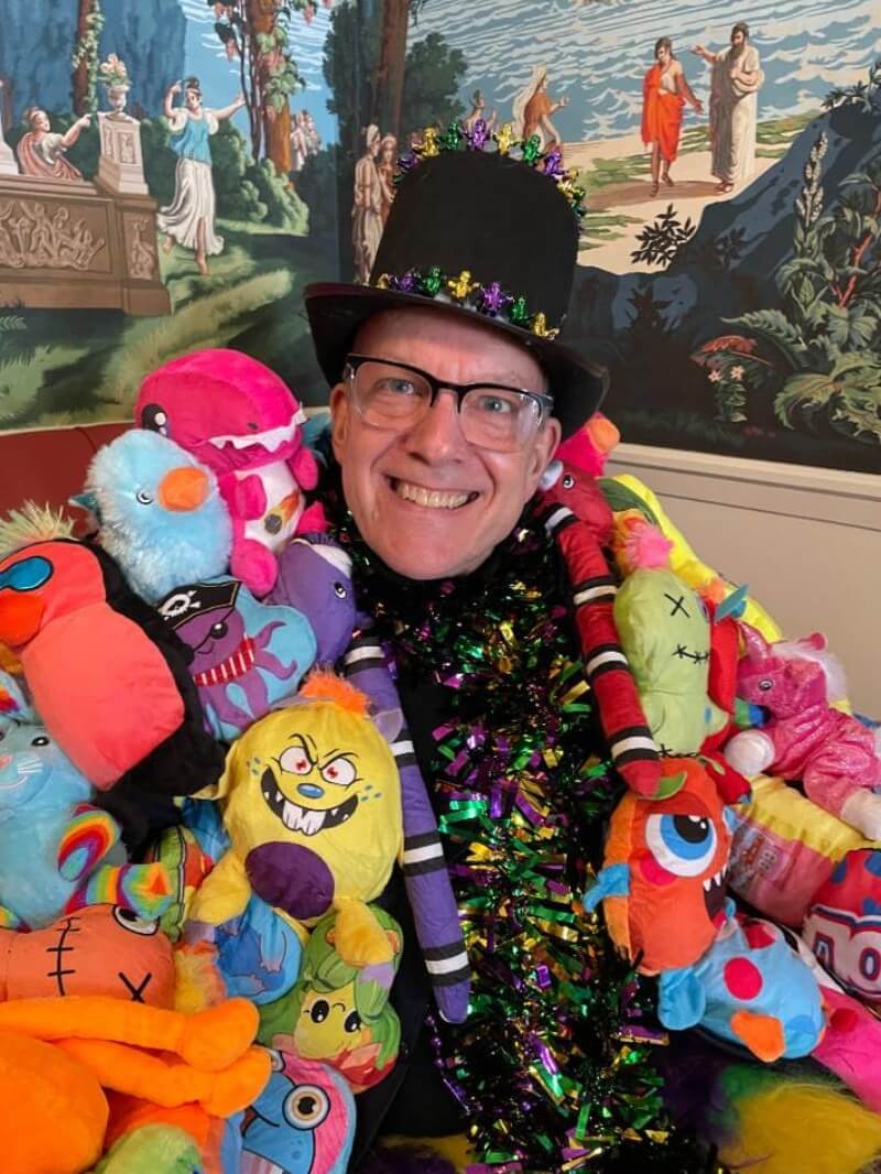 A medium close-up of a middle-aged, white man with horn-rimmed, half-frame glasses smiles for the camera. He is dressed in an elaborate Mardi Gras costume, and based on the angle of his body and how close he is to the corner of the room, it appears that he is sitting for this photo. His bulky but soft jacket is covered entirely with stuffed animals in every color, and it's open to reveal a shirt or wide scarf made of purple, green, and yellow metallic tinsel. He is wearing a black felt top hat decorated with a band of metallic purple, green, and yellow beads. Behind him on the upper half of the adjoining walls is a mural of what looks like a Greek pastoral scene rendered in a slightly desaturated illustration style.