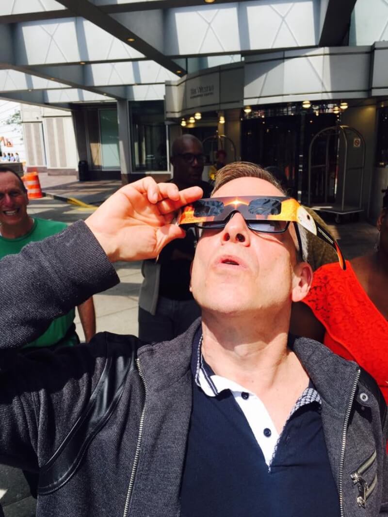 A white man with short, light-colored skin and black glasses is standing outside under or right next to the awning of a hotel. Behind him in the shadows are other people looking off at something or staring at him and smiling. He is holding a pair of disposable eclipse glasses over his own glasses and looking up at the sky with a look of focused effort. He wears a blue polo shirt with a white placket, and over that he is wearing an opened, dark gray, long-sleeved jacket with a zipper front.
