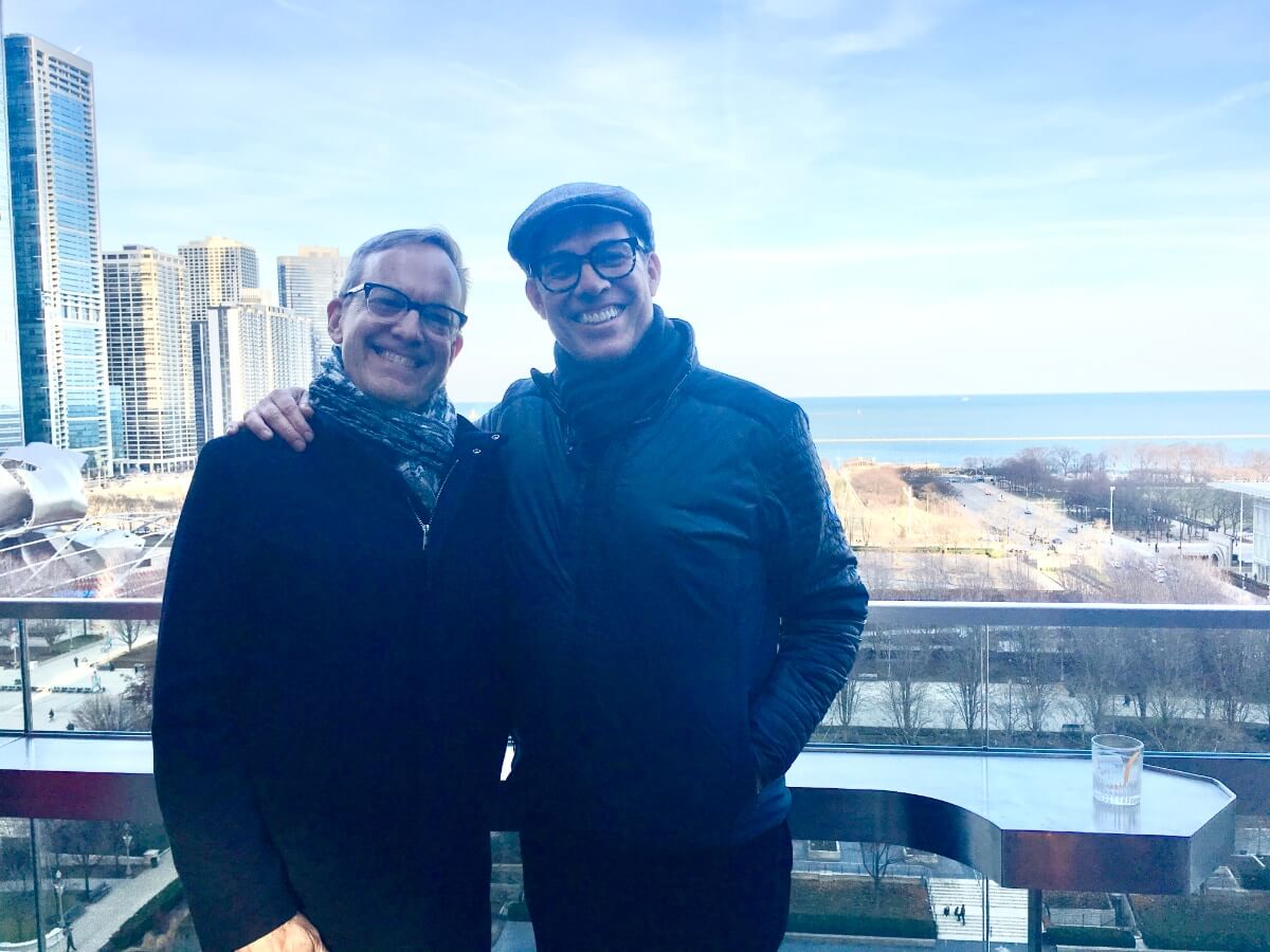 Two men smiling and posing together at what appears to be an outdoor bar or event space on the upper floor of a building. Behind and below them is what appears to be a park, and further back, a large lake or an ocean. It is winter. The park is yellow and and sky is a faint blue that fades to white on the horizon. The men are bundled in dark coats and scarves. The man on the left is white with short, lightly colored hair, and he wears black framed glasses. The man on the right is slightly taller and has his arm around the first man's shoulders . He, too, has black framed glasses, and is wearing a flat cap hat. There is a bluish tinge to the overall photo.