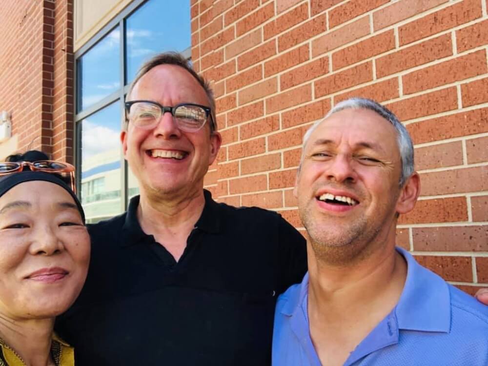 Three adults pose together for the camera, smiling. They are outside next to the red brick wall of an unidentified building. On the left is an asian woman with a tight fitting black cap or black headband that obscures her hair, and pushed up over this head covering are a pair of tortoise-shell oval sunglasses. In the center is a white man with black horn-rimmed, half-frame glasses, wearing a black polo-shirt with the top button undone. On the right is a latino man with dark stubble short, white and gray hair. He is wearing a blue button-down shirt that is open at the top to form a v-shape as it extends out of frame.