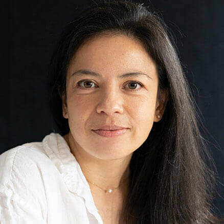 Headshot of Mary Prescott. A medium-light complexioned woman with black hair in an up-do smiles broadly. She is wearing a light floral top, and olive green overalls, standing with her arms crossed in front of a colorful projection.