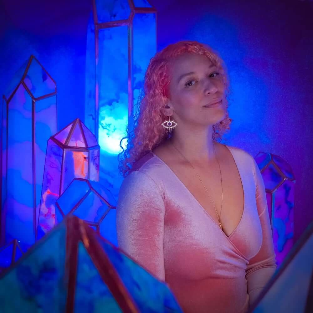 Breanna Thompson An image of a light-skinned woman with pink dyed hair wearing a pink velvet dress, a long gold chain necklace, and one gold earring in the shape of an eye. She is surrounded by large, multi-colored crystals lit in shades of blue.