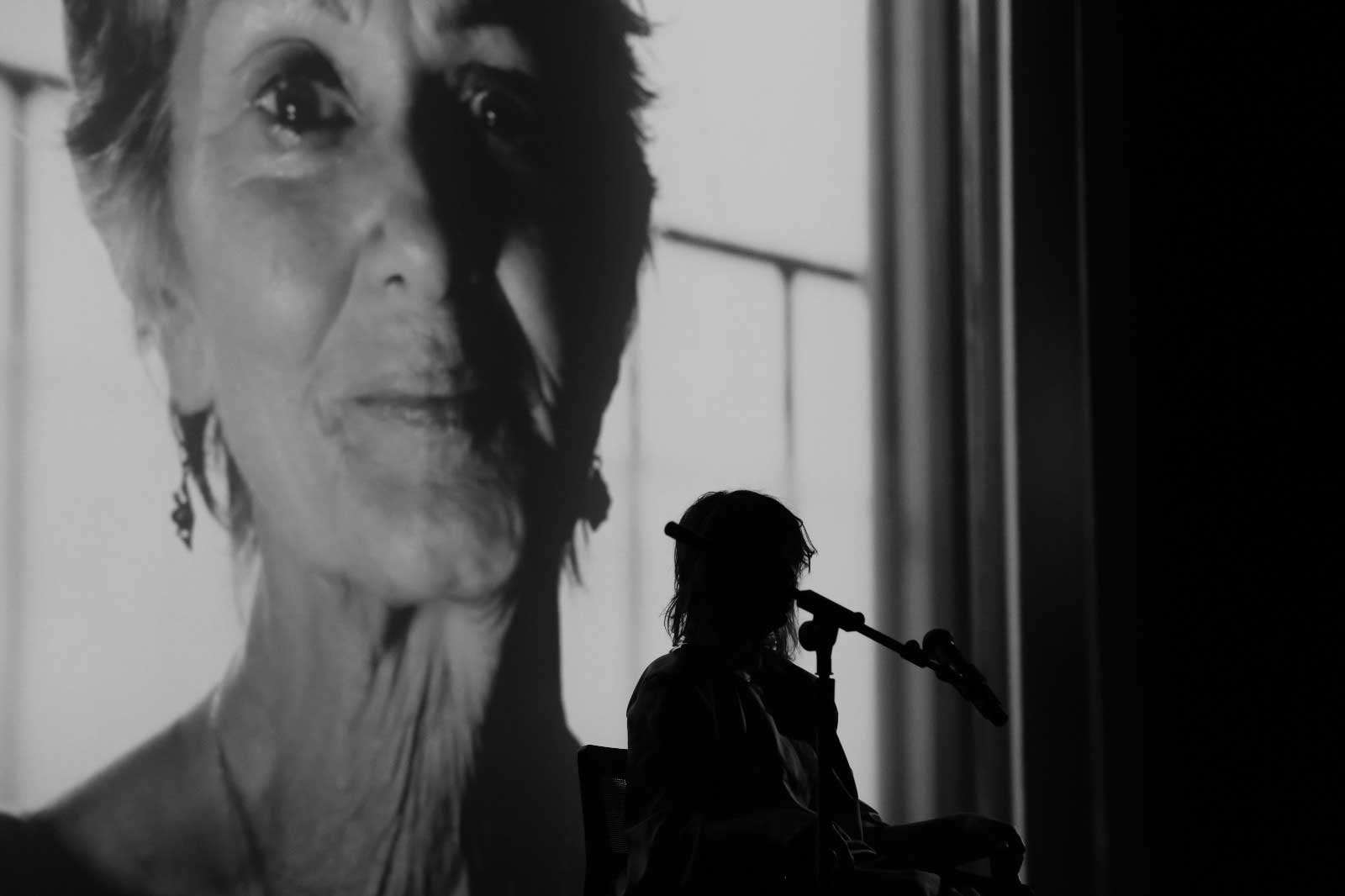 A Latin woman, medium-skinned, seated in a chair in front of a microphone. In the back a big screen, projecting a mid-age Latin woman with a melancholic face.