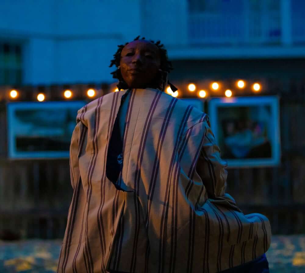 Caleb Dowden An image of a medium-skinned woman sitting down with her back to the viewer. She is wearing a mask tied to the back of her head and a tan blazer with purple stripes; her hairstyle is short locs.