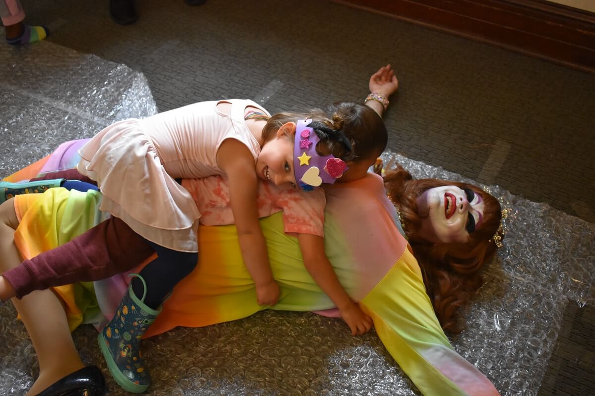 Carla Rossi lies on her back on a sheet of bubble wrap. She is wearing a wig of long red hair and a billowy dress in horizontal peach and yellow stripes. Her arms are spread out and her left leg is folded back in a stylized pose of someone who has fallen, and her face is heavily made up in a stylized bright white base with large purple eyebrow arches and bright red lipstick. She is smiling. Lying on top of her are two children who are approximately four years old. The first is lying face down on Ms. Rossi's stomach and chest. The second is lying on top of the first and has her head turned in the direction of the camera as she laughs with Ms. Rossi.