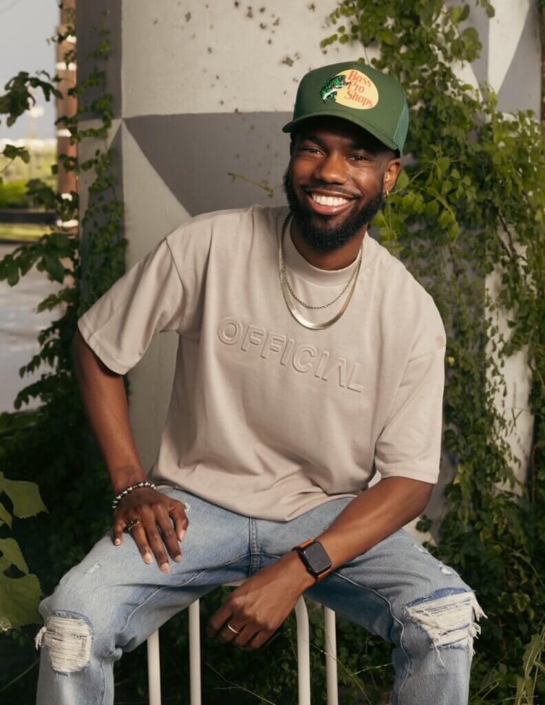 Chris Jones This is an image of a dark-skinned African American man with a full beard. He wears an olive-green baseball cap with a Bass Pro Shop logo, a tan shirt, blue jeans, and fashionable jewelry. He sits on a stool and looks at the camera, smiling broadly at the viewer.