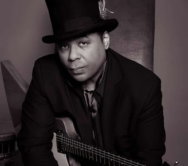 Chris Thomas King A black and white studio portrait of a medium-skinned African American man holding a Gibson Explorer guitar. He is wearing a black tophat with a feather in the hatband and a black suit jacket with a dark-colored satin tie.