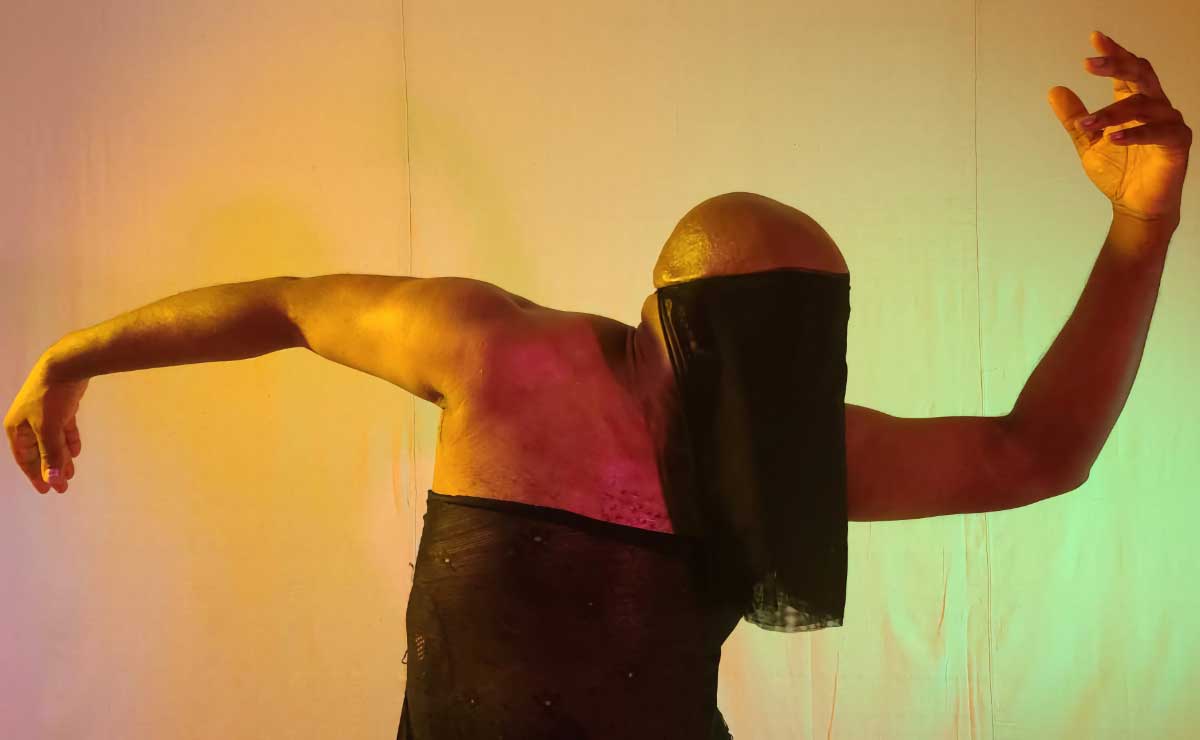A bald Black male-appearing dancer, wearing a sheer, strapless black gown and a long black mourning veil over the face, is turned to the left and slightly bent over, with arms outstretched in opposite directions, one arm angled up and the turned with the palm facing down, so that together the arms form a tilde-shaped curve.