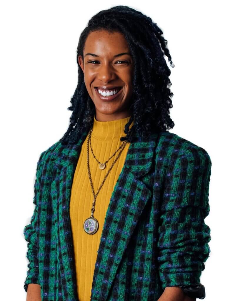Deja M. Jones An image of a medium light-skinned, Black woman with dark brown locs wearing a yellow shirt, blue and green plaid blazer, and locket jewelry around her neck.