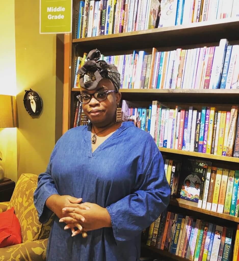 Desiree S. Evans An image of a medium-brown-skinned Black woman standing by a bookshelf smiles at the camera. She is wearing black-framed eyeglasses, a long denim shirt, large brown spiral earrings, a necklace with a crystal, and a brown-and-white patterned head wrap.
